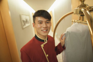 左手でものに触れながら赤い制服を着て歯を見せて笑うホテルのボーイ